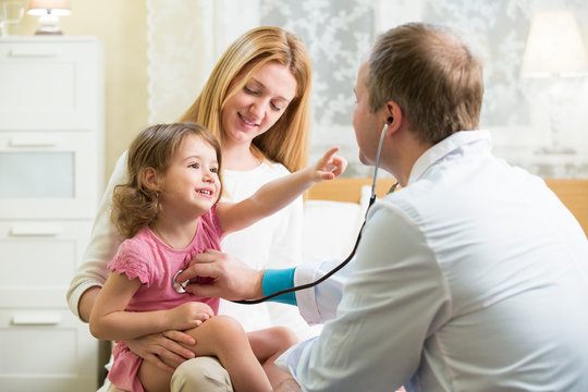Male Pediatrician examining cute little girl with stethoscope. Kid happily smiling and pointing on doctor. Mother holding her kid. Doctor visit his patient at home.