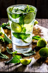 Glass of alcohol cocktail or lemonade. Mohito cocktail with lemon, lime, mint, ise cubes. Cold drink. Fresh lime and mint on wooden background