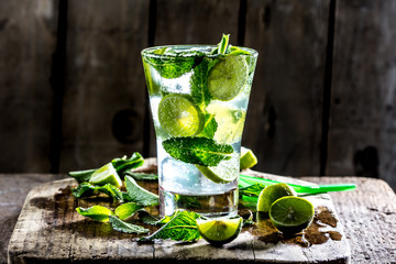 Glass of alcohol cocktail or lemonade. Mohito cocktail with lemon, lime, mint, ise cubes. Cold drink. Fresh lime and mint on wooden background
