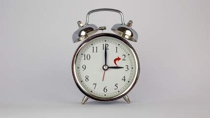 Silberner Wecker vor hellem Hintergrund mit rotem Pfeil zur Zeitumstellung