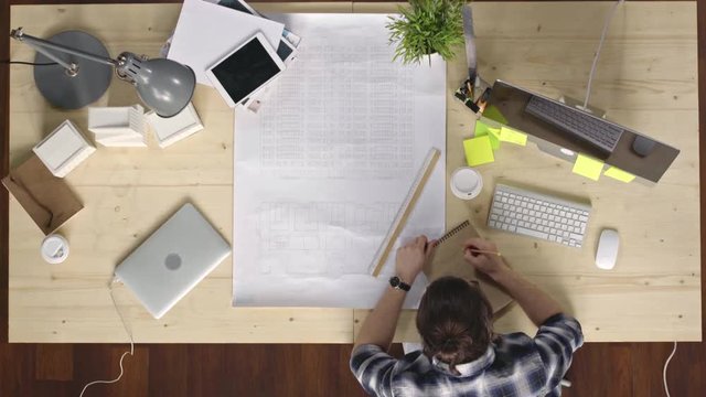 Top view of man sitting at office table with building plan on it and drawing chart in sketchbook with pencil then taking typing on a computer and drinking coffee from disposal cup