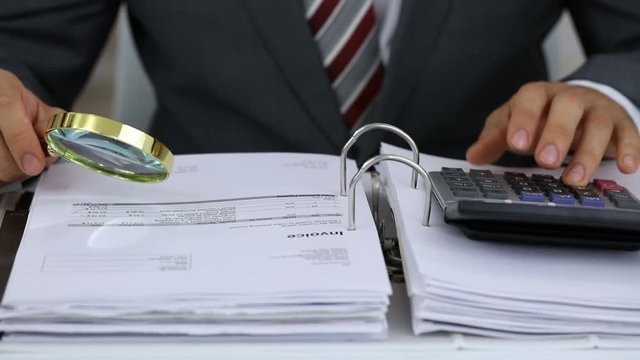 Close-up Of A Businessman Holding Magnifying Glass Calculating Invoices Using Calculator