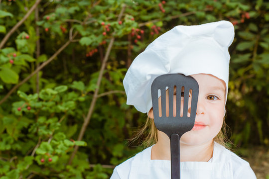 Девочка-поваренок закрывает один глаз лопаткой для жарки