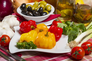 Pickled peppers, red and yellow, five pieces on a white plate next to olives, olive oil, garlic and cherry tomatoes.