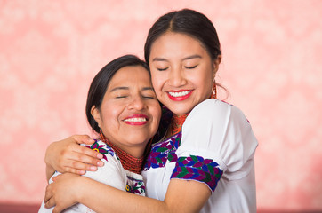 Beautiful hispanic mother and daughter wearing traditional andean clothing, embracing while posing happily together interacting for camera, pink studio background