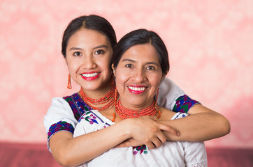 Beautiful hispanic mother and daughter wearing traditional andean clothing, embracing while posing happily together interacting for camera, pink studio background