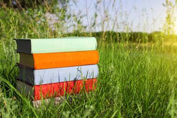 Stack of multicolored books on the green grass on the background of beautiful nature surrounded by meadows at sunny day.Library at outdoor. Back to school.