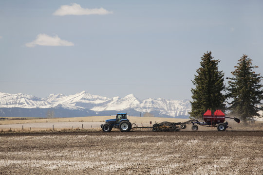 Tractor with air seeder planting crop in field with snow covered mountains and blue sky and clouds,Alberta Canada
