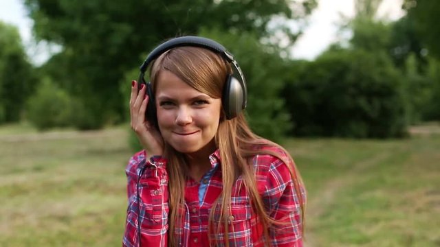 Young attractive girl with headphones bursts the bubble of chewing gum. Girl listening to music in a summer park. She smiles.