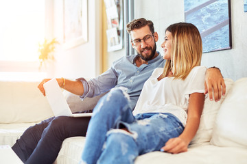 Young Couple Sitting On Sofa Using Laptop looking at each other and smiling