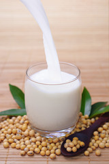 Pouring soy milk into clear glass decorated by green leaf and soybean on wooden spoon with bamboo background.