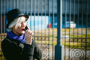 portrait of young beautiful woman with white hair, in a black coat, a skirt and a black hat, smoking an electronic cigarette, blowing the smoke vapor