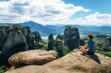 Young woman sits back to the camera on top of the mount and looks at the valley and rocks. Concept of travelling, happiness, freedom. Traveler enjoying the mountain landscape of Meteora, Greece