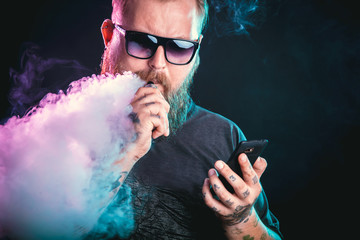 Men with beard  in sunglasses vaping and releases a cloud of vapor holding a mobile phone