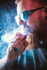 Men with beard  in sunglasses vaping and releases a cloud of vapor.