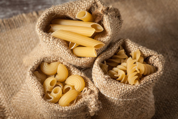 Various mix of pasta on wooden rustic background, sack and wooden spoons. Diet and food concept.