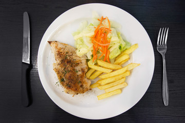 Chicken steak with vegetable and french fries on a white plate on black wooden background (top view).