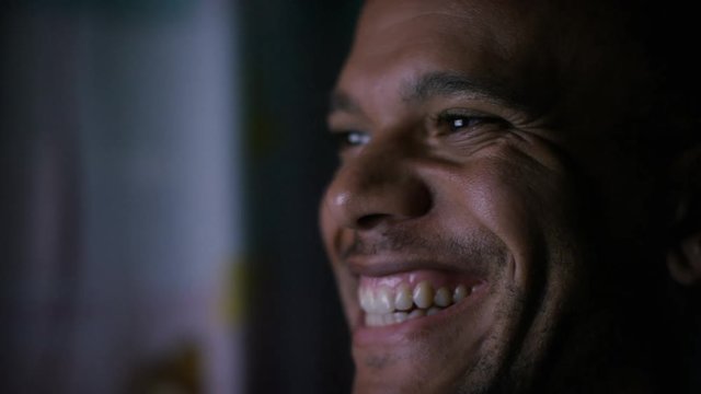 Man reacts with laughter at something he is watching on a screen at night in the dark
