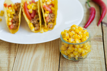 Yellow corn kernels in a small glass bowl, chili peppers and tacos with chicken meat and tomato on wooden background