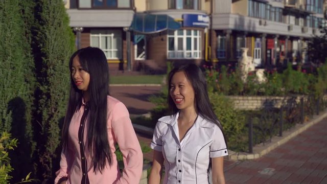 Young Friends Walking On The Street. On The Background Urban View With Buildings, Shops, Cafe. Two Women Talking And Walking On The Pavement. Asian Girls Wearing In Casual Shirts At Sunny Day.