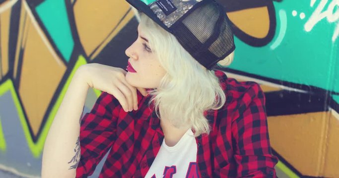 Beautiful young hipster blond woman wearing a baseball cap with a pierced lower lip sitting in front of colorful graffiti looking to the side with a serious expression.