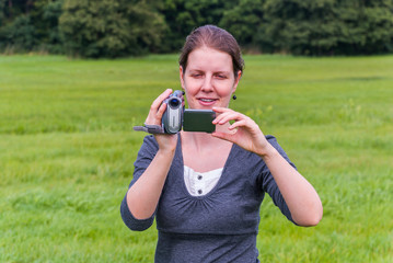 Woman with brown hair practising filming with her retro camcorder, standing on a meadow during...