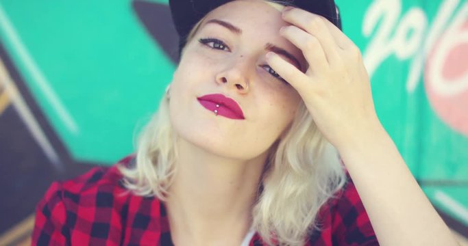 Gorgeous young blond woman with a pierced lip wearing a trendy baseball cap and crimson lipstick posing for the camera in front of colorful graffiti