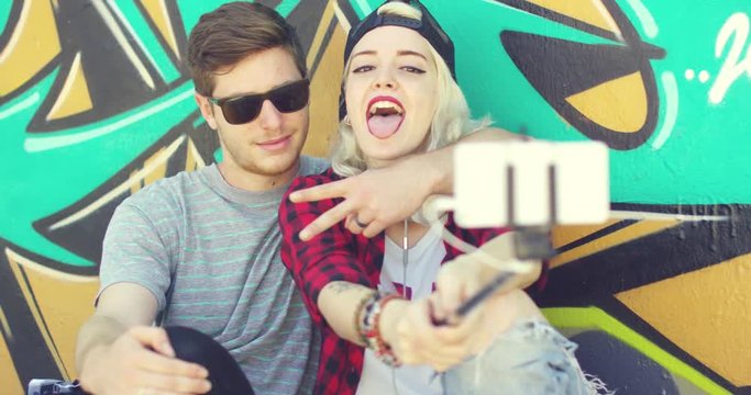Modern fashionable young couple taking a selfie as they pose sitting together in front of a colorful graffiti covered wall