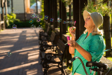 Senior woman blowing bubbles. Lady in hat outdoors. Return to childhood. The lightness of breath.