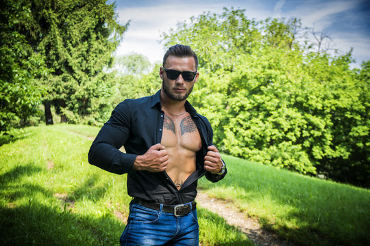 Handsome Hunk Man Outdoor in City Park, Opening his Shirt to Show Muscular Torso and Chest During Daytime, Wearing Black Shirt and Sunglasses