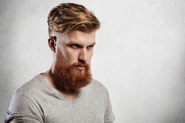 Portrait of attractive bearded young Caucasian man in casual gray t-shirt standing against studio wall background with copy space for your text or advertising content, looking serious and confident