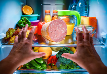 Human hands reaching for food at night in the open refrigerator - obrazy, fototapety, plakaty