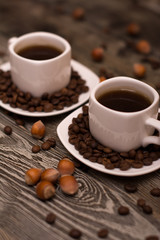 two small white cups of coffee with cocoa beans, slices of chocolate and hazelnuts on wooden background