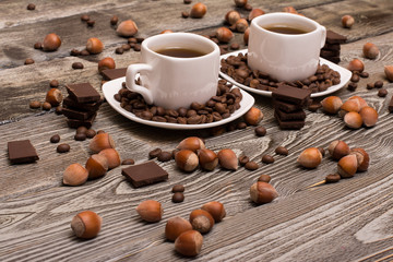 two small white cups of coffee with cocoa beans, slices of chocolate and hazelnuts on wooden background