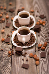 Obraz na płótnie Canvas two small white cups of coffee with cocoa beans, slices of chocolate and hazelnuts on wooden background