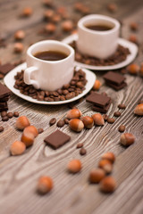 two small white cups of coffee with cocoa beans, slices of chocolate and hazelnuts on wooden background