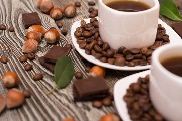 two small white cups of coffee with cocoa beans, slices of chocolate, hazelnuts and green leaves on wooden background