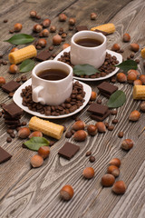 two small white cups of coffee with, cookies, cocoa beans, slices of chocolate, hazelnuts and green leaves on wooden background