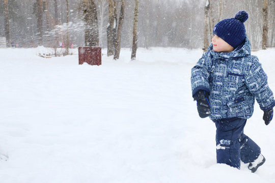 Happy little boy throws snow in park during snowfall in winter d