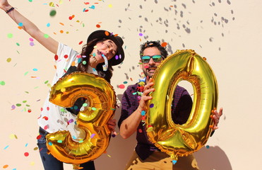 Cheerful couple celebrates a thirty years birthday with big golden balloons and colorful little pieces of paper in the air.