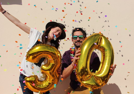 Cheerful couple celebrates a thirty years birthday with big golden balloons and colorful little pieces of paper in the air.