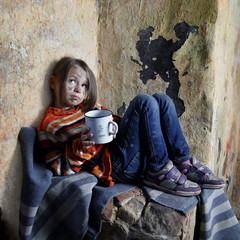 Sad little girl with dirty face sits on old blanket in basement with iron mug in hands - refugee...