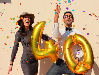 Cheerful couple celebrates a forty years birthday with big golden balloons and colorful little pieces of paper in the air.