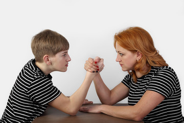 Redhead woman and teenage boy in similar t-shirts fight on hands on gray background - mother and son arm wrestling - solution of disagreements in the family