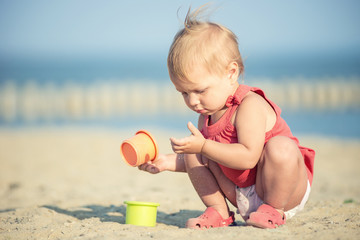 Baby playing on the sandy beach near the sea. Cute little girl in red dress with sand on tropical beach. Ocean coast.