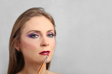 Pretty young woman with bright evening makeup applies lipstick using brush on gray background with blank copy space