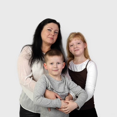 Brunette adult woman and teenage blonde girl embrace little boy smiling on gray background in square - mother, son and daughter - family relations and love concept