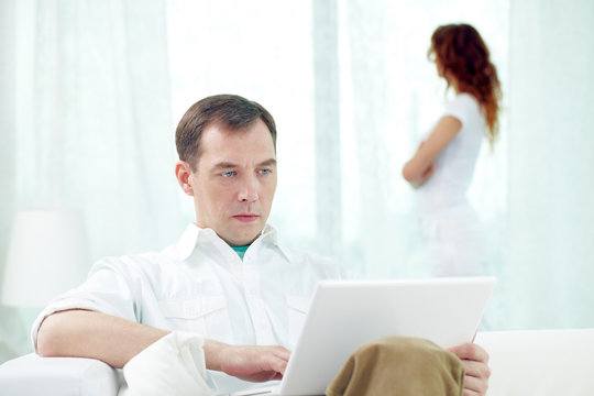 Young man sitting with laptop with woman standing near the window on the background