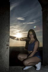 Chica morena a contraluz con el sol por detrás en lo alto de la torre en la montaña al atardecer. Chica joven posando. Sesión con modelo en el castillo.