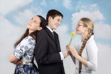 Teenage boy stands back to back with one girl and gives flowers to another on cloudy blue sky background - Inconstancy and volatility of youth concept